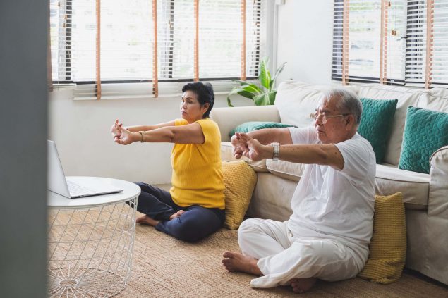 An older Asian couple sits on the floor doing stretching exercises with their hands and arms.