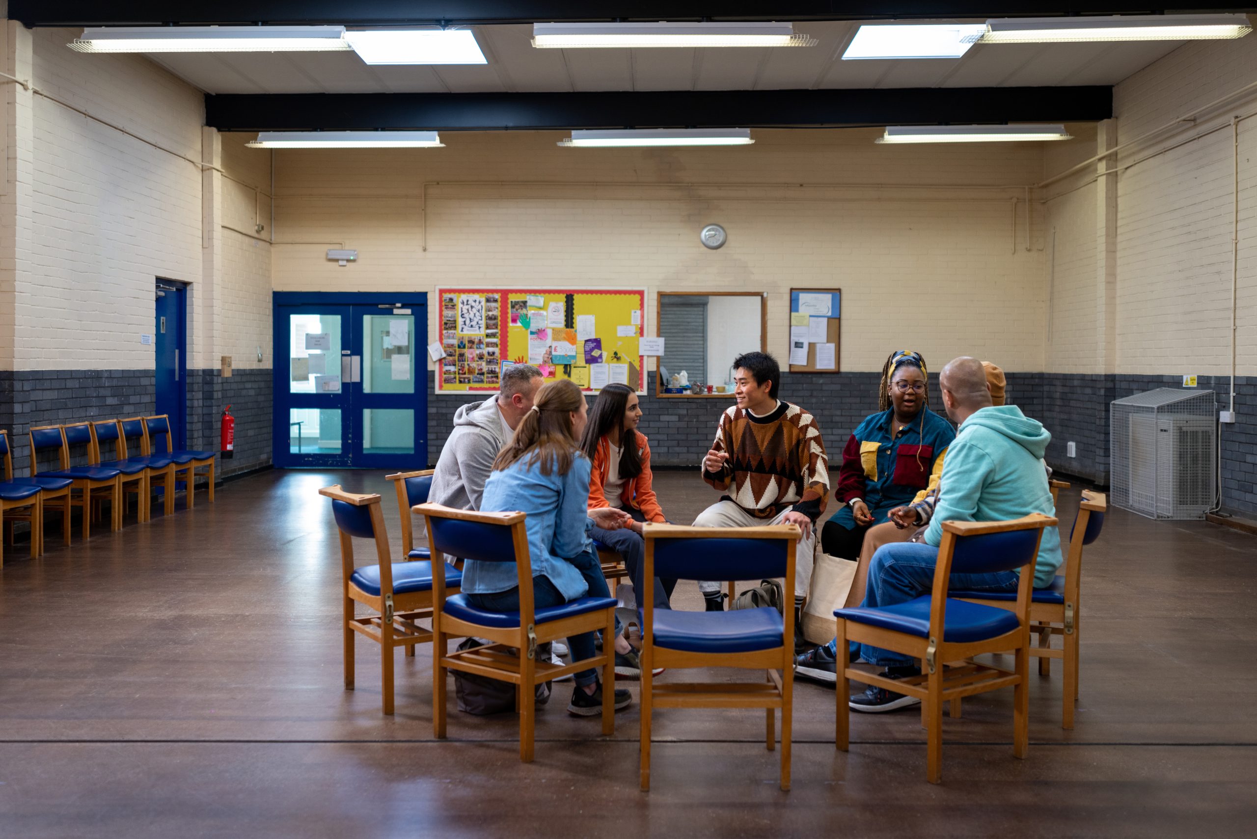 A multi racial group of people meeting; they sit in blue chairs arranged in a circle facing each other