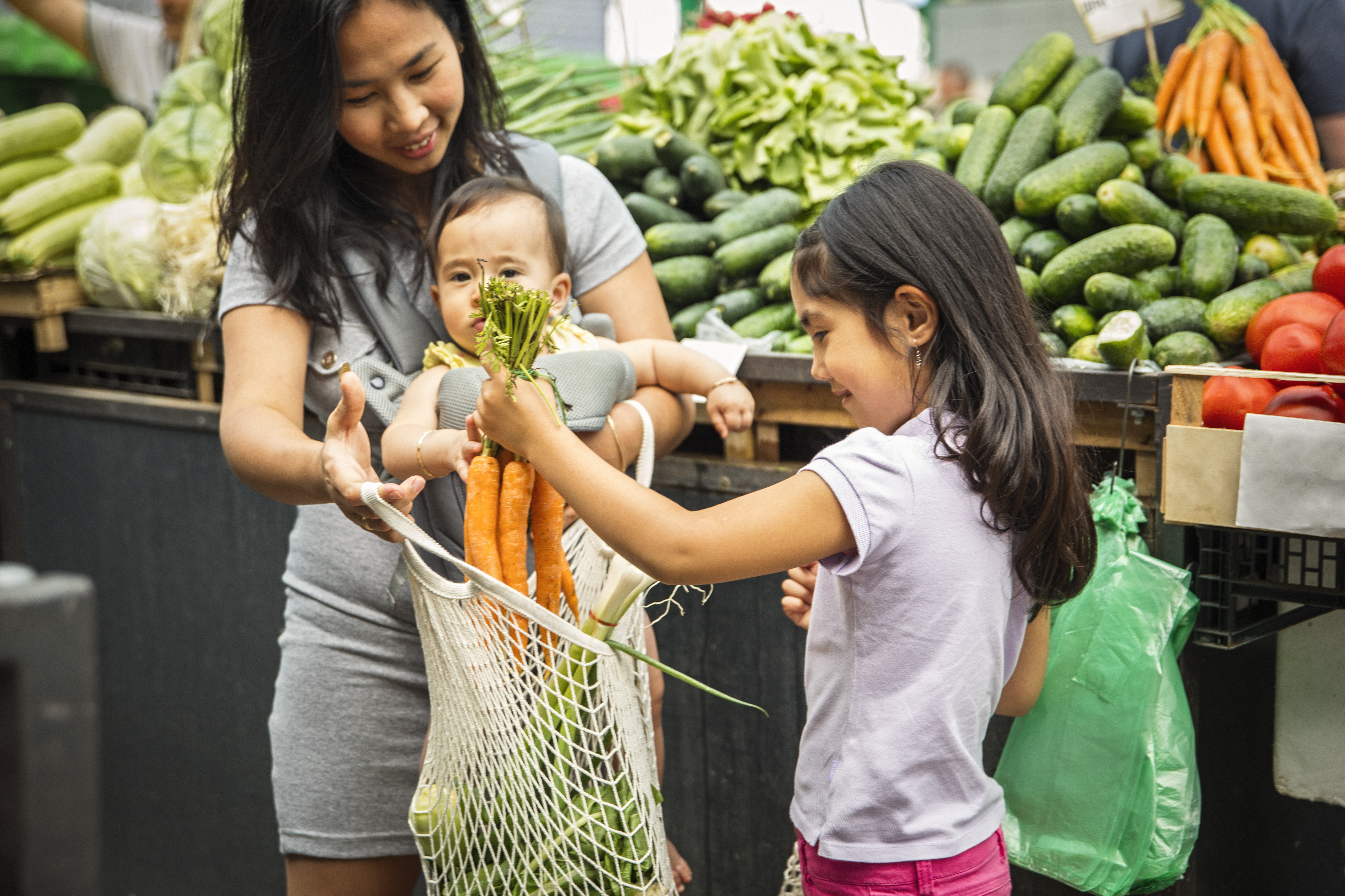 A mother and her two children placing vegetables in a mesh grocery bag