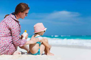 A woman applies sunscreen to the shoulders and back of a young child as they sit on a beach.