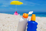 Two bottles of sunscreen sit upright in the sand of a beach, with the ocean in the background.